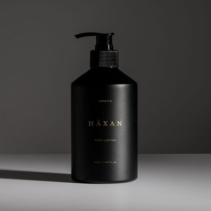 A matte black pump bottle HÄXAN body lotion, featuring gold lettering, set against a dark gray studio background with a soft shadow.