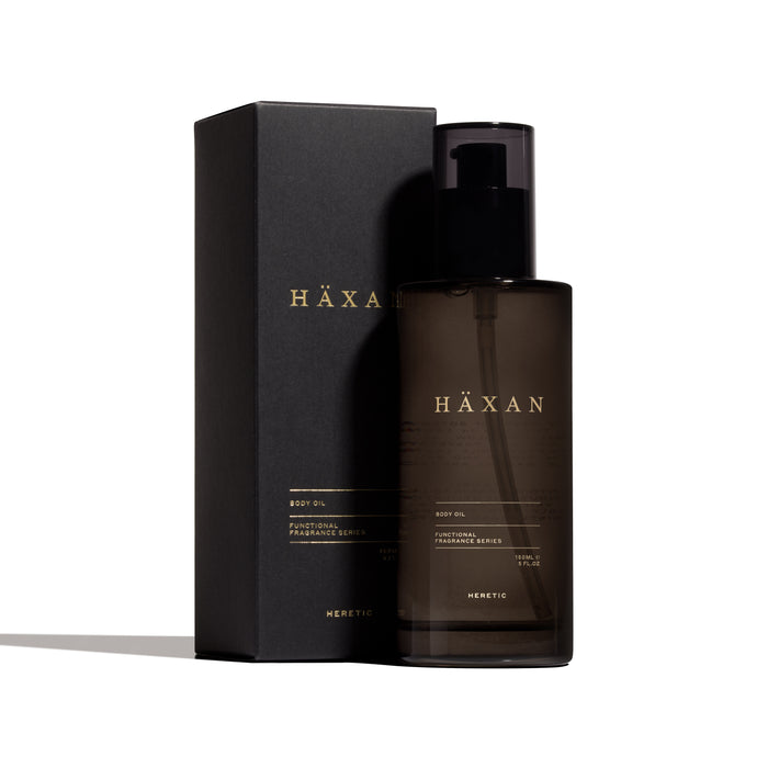 A bottle of HÄXAN body oil next to its black packaging box, both featuring gold lettering, on a clean white background.